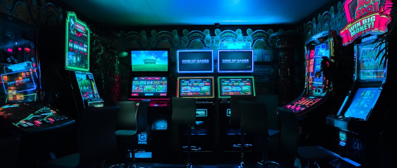 Dimly lit gaming arcade featuring a variety of brightly illuminated slot machines and video gaming screens. Neon signs and colorful displays create a vibrant atmosphere. The arrangement of gaming machines suggests an inviting space for entertainment, with chairs positioned for players.