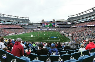 A large sports stadium filled with spectators. The field is green with white markings, and players in red and white uniforms are actively engaged in a game. The stands are packed with people wearing various colored clothing, and some sections of the crowd are more densely populated. Digital screens and advertisements are visible around the stadium, adding to the vibrant atmosphere.