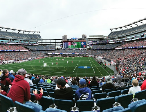 A large sports stadium filled with spectators. The field is green with white markings, and players in red and white uniforms are actively engaged in a game. The stands are packed with people wearing various colored clothing, and some sections of the crowd are more densely populated. Digital screens and advertisements are visible around the stadium, adding to the vibrant atmosphere.