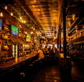 A dimly lit bar interior with a warm ambiance, featuring several television screens displaying sports broadcasts above a wooden counter. Patrons are seated at the bar and tables, engaged in conversation. The ceiling is ornate with decorative panels and hanging light fixtures add a cozy glow to the scene.