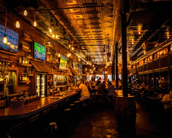 A dimly lit bar interior with a warm ambiance, featuring several television screens displaying sports broadcasts above a wooden counter. Patrons are seated at the bar and tables, engaged in conversation. The ceiling is ornate with decorative panels and hanging light fixtures add a cozy glow to the scene.