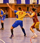 A dynamic indoor sports scene with players in motion on a court. Two athletes in blue and yellow uniforms are engaged in an intense handball game, with one player gripping the other's jersey. The background shows other players, several seated people, and advertisements on the walls.