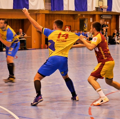 A dynamic indoor sports scene with players in motion on a court. Two athletes in blue and yellow uniforms are engaged in an intense handball game, with one player gripping the other's jersey. The background shows other players, several seated people, and advertisements on the walls.