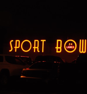 The image depicts a brightly lit neon sign that reads 'SPORT BOWL' with an additional sign indicating '24 LANES'. The scene appears to be taken at night, with several vehicles parked in front of the establishment.
