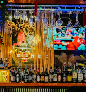 An illuminated bar counter features a variety of liquor bottles, with colorful neon lights adding vibrancy. Glasses hang upside down above the bar, and a large television screen in the background displays a sports event.