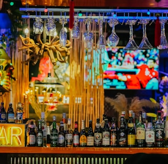 An illuminated bar counter features a variety of liquor bottles, with colorful neon lights adding vibrancy. Glasses hang upside down above the bar, and a large television screen in the background displays a sports event.