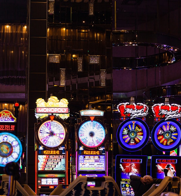 A lively casino scene featuring a row of bright, colorful slot machines with themes like Monopoly, Michael Jackson, and Lord of the Rings. The gaming machines display a variety of flashing lights and digital screens, creating an energetic and exciting atmosphere. Reflections of the lights are visible in glossy surfaces above the machines.