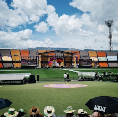A sports stadium with a large stage set up on the field, hosting an outdoor event. Spectators wearing colorful hats sit in the foreground. The seating areas display vibrant colors, and a clear blue sky with scattered clouds is visible. Large screens display graphics related to the event.