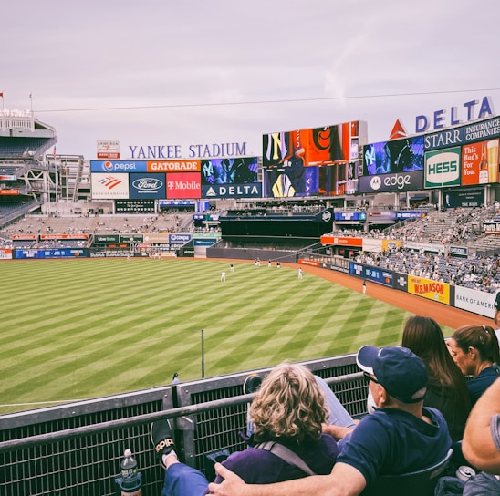 A baseball stadium filled with spectators watching a game on a neatly maintained grass field. The stadium features large advertising screens displaying various logos and brands. Fans sitting in the foreground are dressed casually, enjoying the sporting event.