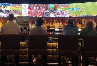 A group of people sit at a bar with their backs to the camera, watching multiple sports games displayed on large screens. The bar counter is lined with various bottles of liquor, and the atmosphere suggests a casual and relaxed social setting.
